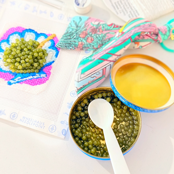 A styled flat-lay of craft supplies on a white surface, including a small embroidered patch with green beads, patterned fabric, printed cards, and two open containers of green seed-like beads with a white spoon.